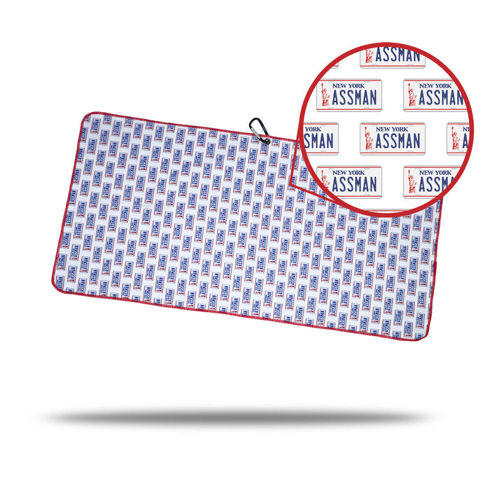 The ASSMAN - Towel displays a repeated pattern of New York “ASSMAN” license plates in red and blue with a red border. It includes a black cord, handy clip, eyelet for easy hanging or storage, and features a circular close-up inset.