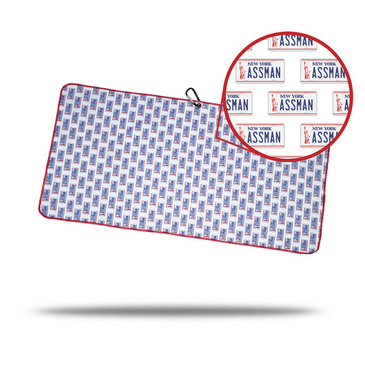 The ASSMAN - Towel displays a repeated pattern of New York “ASSMAN” license plates in red and blue with a red border. It includes a black cord, handy clip, eyelet for easy hanging or storage, and features a circular close-up inset.