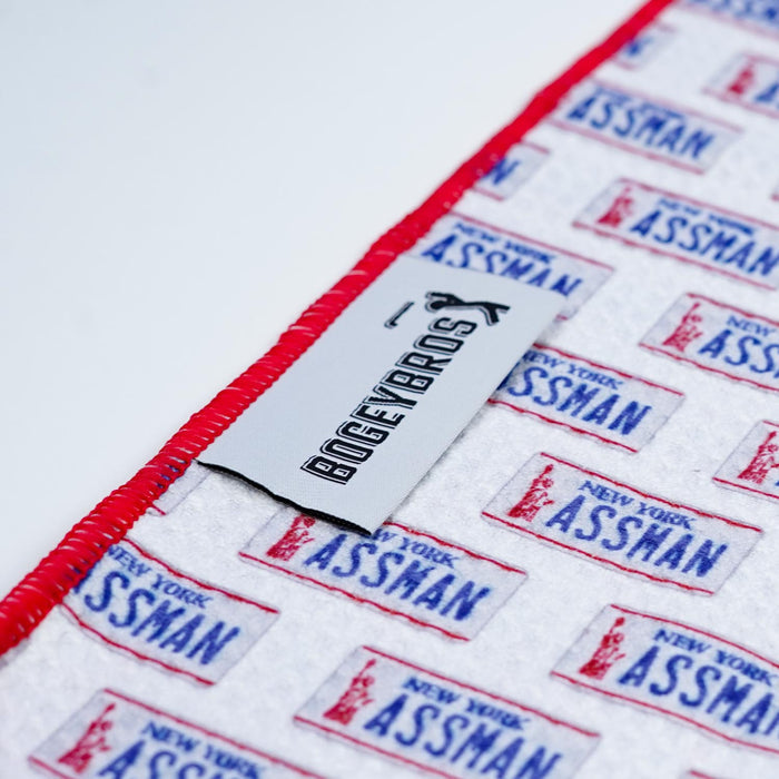 A close-up of the ASSMAN - Towel, featuring white microfiber with repeating red and blue “NEW YORK ASSMAN” license plates. A white Bogey Bros tag is sewn by the red-stitched edge on a white background.