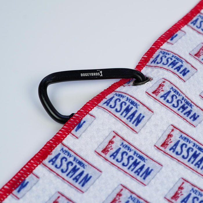 The ASSMAN - Towel is a white microfiber towel with red trim, featuring double-sided blue and red NEW YORK ASSMAN license plate prints. It includes a black BOGEYBROS carabiner clip and eyelet on the corner, displayed on a plain white background.