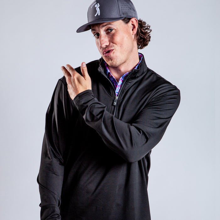 A person with long curly hair wearing a gray baseball cap and a Black - Q-Zip stands against a plain light background, pointing sideways and making a playful, exaggerated facial expression.