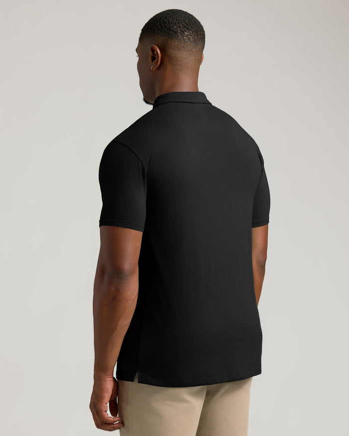 A man viewed from the back wearing a black short-sleeve polo shirt and beige trousers, photographed against a plain light gray studio background.