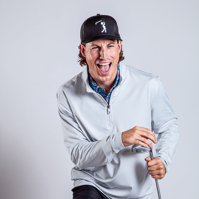 A man in the Bone - Q-Zip, a light gray athletic fit quarter-zip, smiles widely with his tongue out. He wears a black golf cap and patterned shirt, holding a golf club while leaning forward against a plain background.
