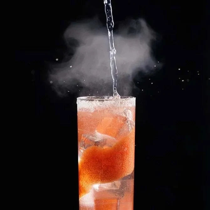 Glass of orange cocktail with ice cubes and liquid being poured, popping smoke bubble against a black background.