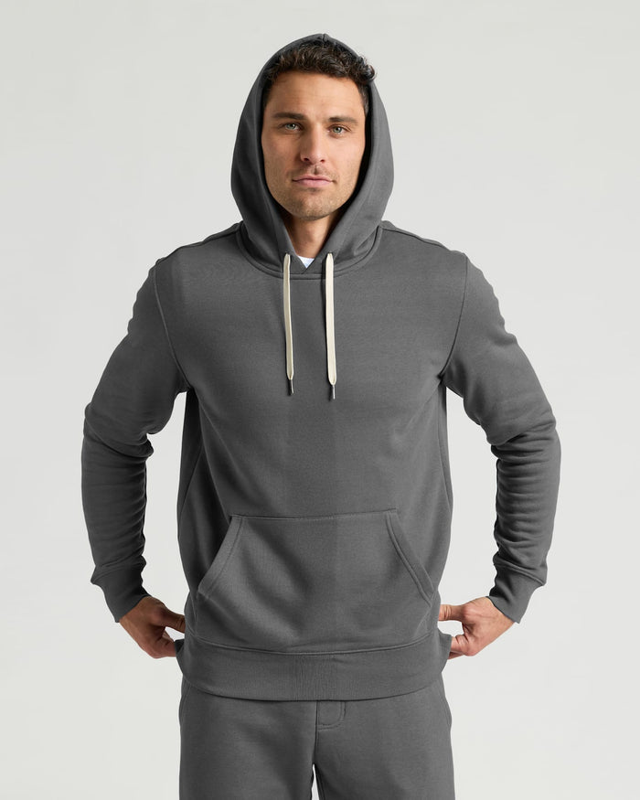 A man wearing a dark gray pullover hoodie with light beige drawstrings and matching gray sweatpants, standing against a light gray studio background.