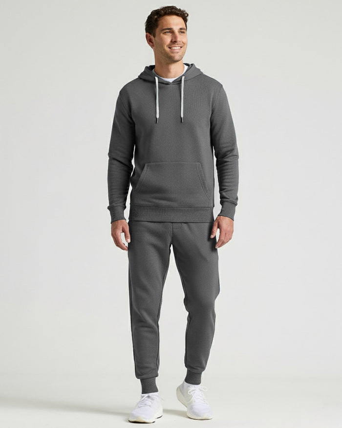 A man modeling a dark gray ribbed hoodie and matching jogger pants set, paired with white sneakers, against a light gray studio background.