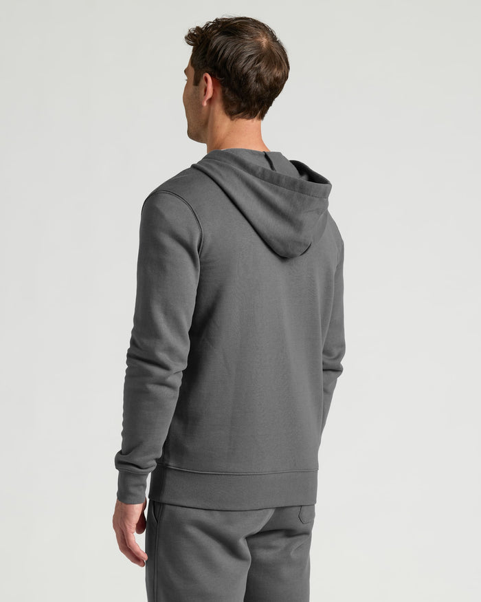 A man wearing a dark gray pullover hoodie and matching sweatpants, shown from a rear three-quarter view against a light gray studio background.
