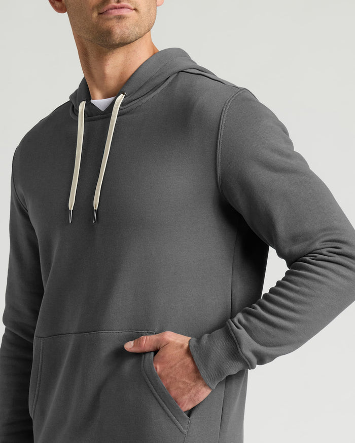 A man wearing a dark gray pullover hoodie with cream-colored drawstrings, shown from the chest up against a light gray studio background.