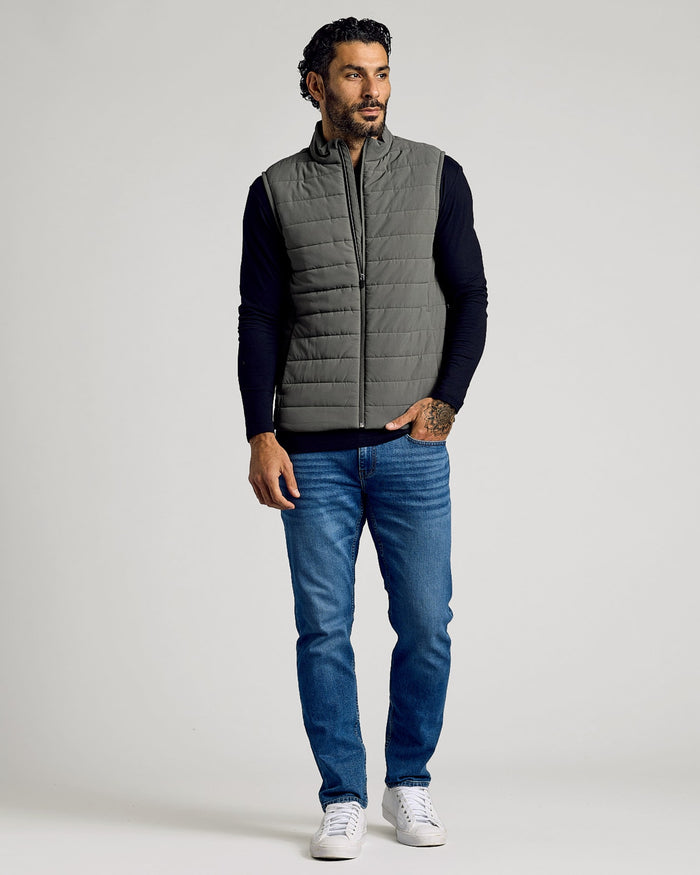 Man wearing a grey Carbon Puffer Vest over a black long-sleeve shirt paired with blue jeans and white sneakers, standing against a plain background.