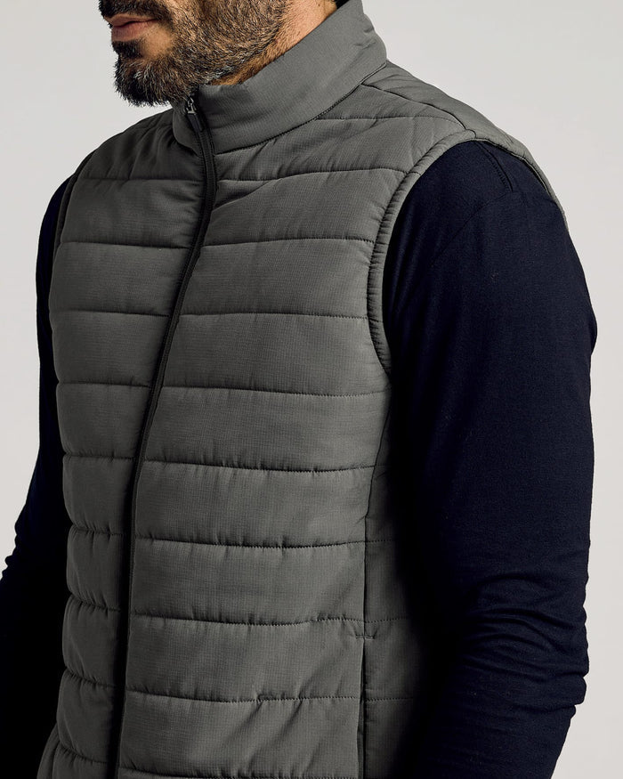 Man wearing a gray Carbon Puffer Vest over a black long-sleeve shirt, featuring a high collar and horizontal quilted stitching.