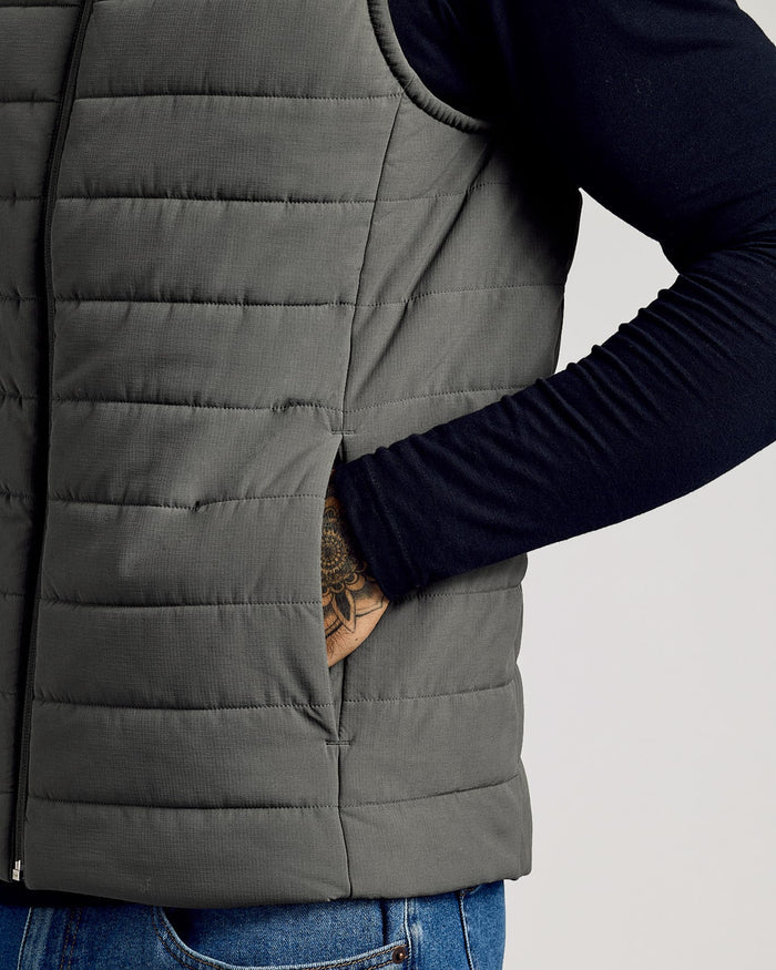 Close-up of a person wearing a charcoal gray Carbon Puffer Vest over a navy long-sleeve shirt with one hand inside the vest pocket, showcasing the vest's quilted texture and casual fit.