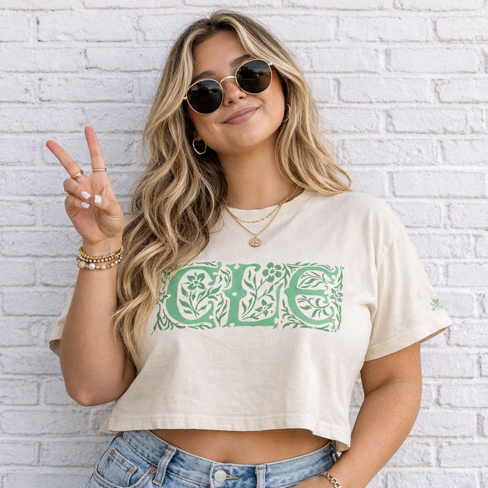 Woman wearing a beige crop top with green floral design and sunglasses, standing against a white brick wall.