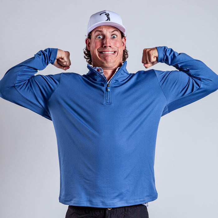 A person in the athletic fit Cooler Blue - Q-Zip flexes both arms and smiles widely with a humorous expression, wearing a white cap and standing against a plain light background.