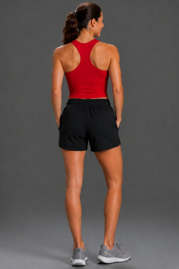 A woman shown from behind wearing a red athletic tank top and black athletic shorts, paired with gray sneakers, against a solid gray studio background. || Black