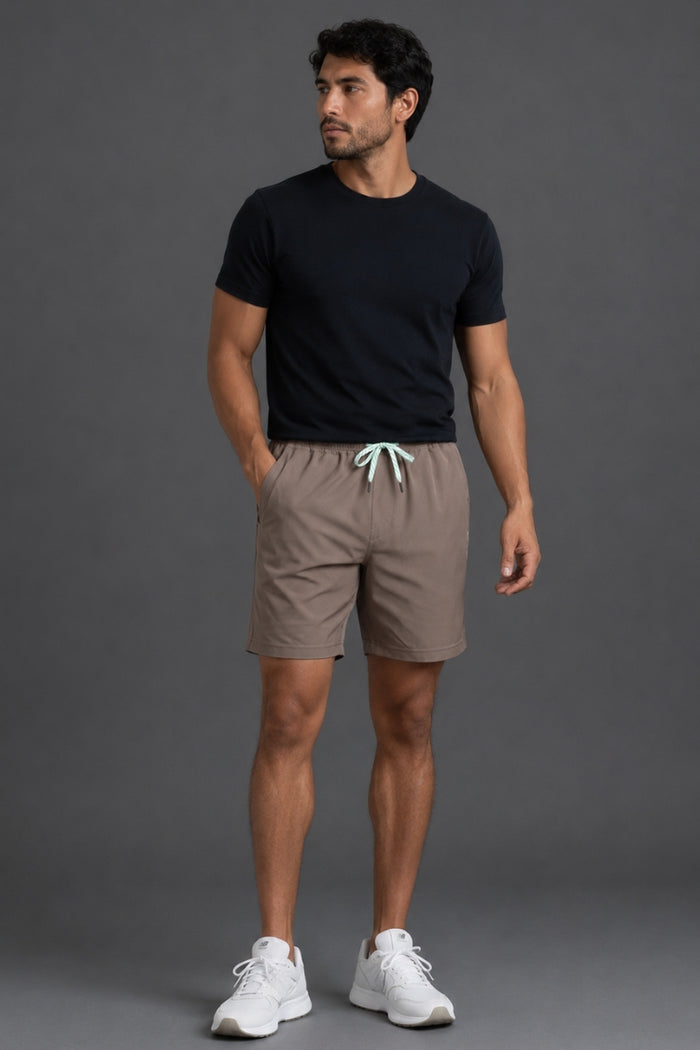 A man wearing taupe-brown athletic shorts with a mint green drawstring, paired with a black t-shirt and white sneakers, shown from the waist down against a gray studio background. || Mocha