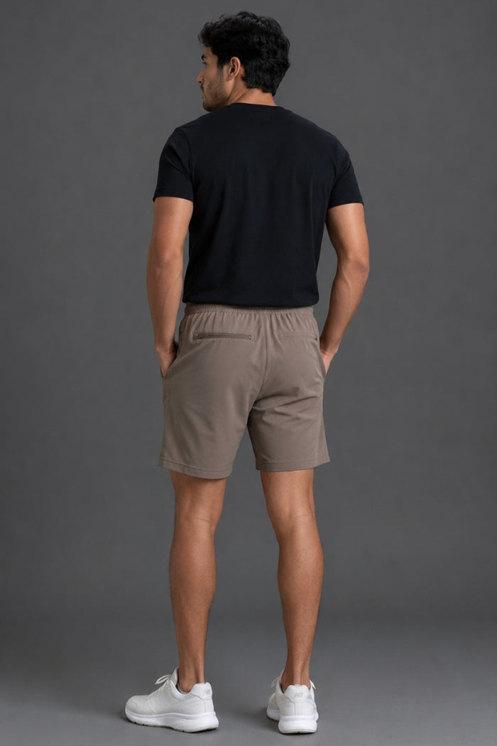 A man standing with his back to the camera, wearing a black short-sleeve t-shirt, beige athletic shorts, and white sneakers against a solid gray studio background. || Mocha