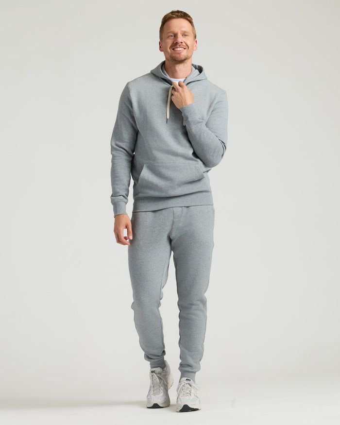 A man wearing a Dark Heather Gray Fleece French Terry Pullover Hoodie paired with matching sweatpants and sneakers, smiling while standing against a plain background.