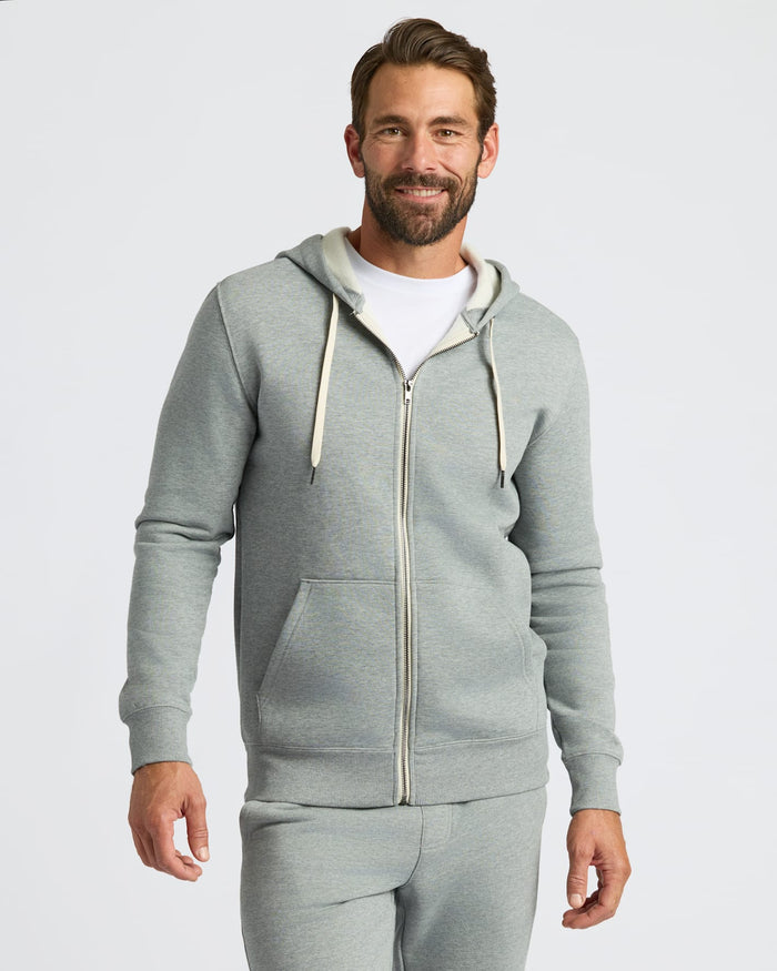 A man wearing a light gray zip-up hoodie with cream drawstrings and matching gray sweatpants, standing against a light gray studio background.