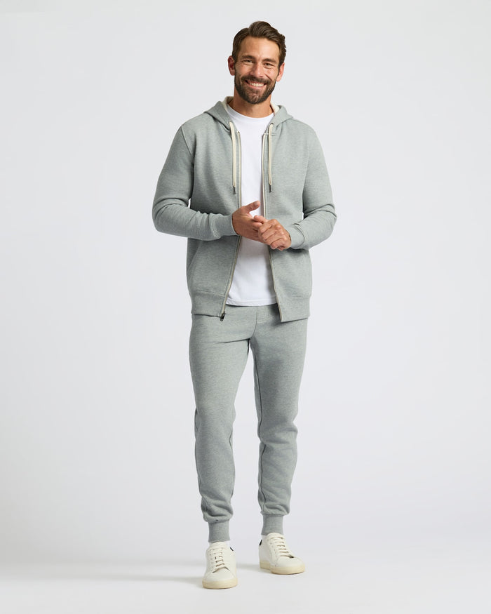 A man modeling a light gray two-piece tracksuit featuring a zip-up hoodie with white drawstrings and matching jogger pants, styled over a white t-shirt and cream sneakers against a light gray studio background.