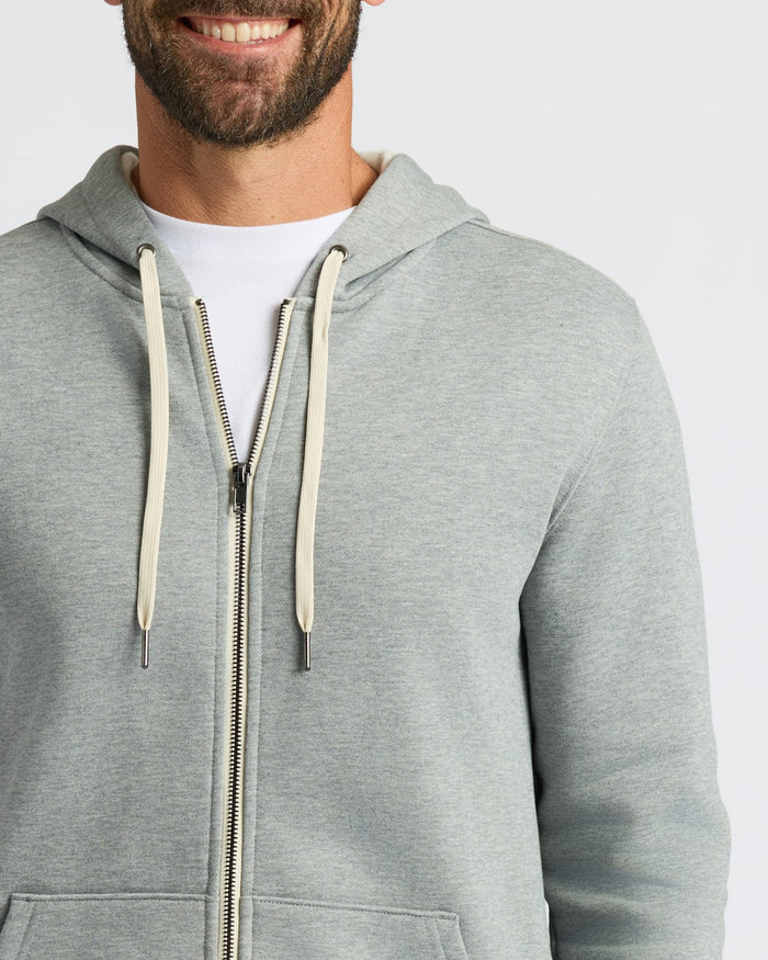 A close-up of a man wearing a light gray heather zip-up hoodie over a white t-shirt, showing the front of the hoodie with its drawstrings and zipper.