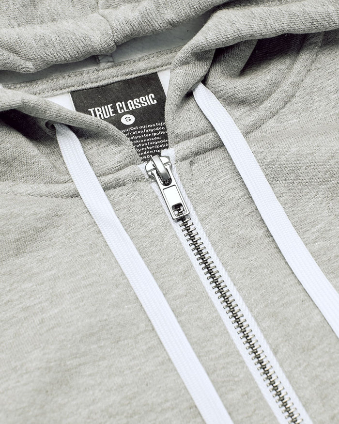 Close-up of a light gray zip-up hoodie with white drawstrings and a black neck label reading TRUE CLASSIC.