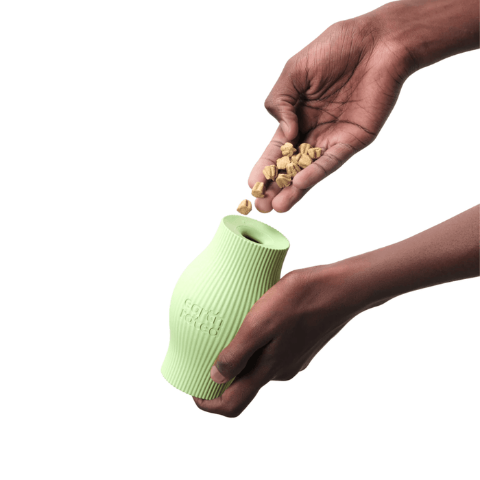 White background with a pair of hands pictured filling a pale green Earth Rated Treat dog toy with dog treats.