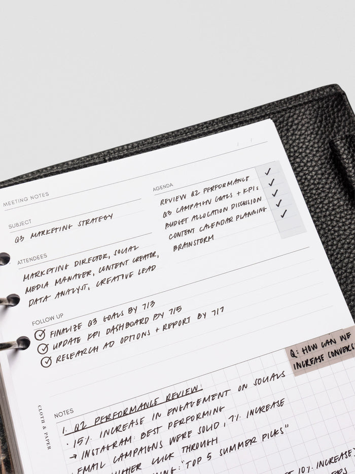 A meeting notes insert filled in with handwritten text, featuring sections for subject, agenda, attendees, follow-up, and a gridded notes area. The page rests in a black leather planner.