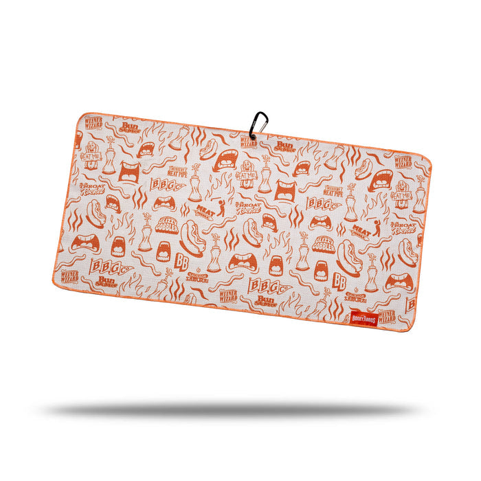 The Glizzy Gobbler - Towel is a rectangular microfiber towel with orange BBQ-themed grills, flames, utensils, and condiments on white. Features double-sided print, orange edging, black hanging loop, red logo tag (bottom right), and shadow below.