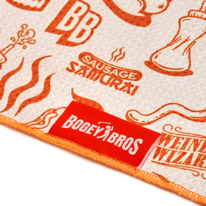 Close-up of the Glizzy Gobbler - Towel, a light-colored microfiber towel with orange cartoon food graphics and text like SAUSAGE SAMURAI and WEINER WIZARD. Features 2-sided print, orange stitching, red BOGEY BROS tag, clip, and eyelet.