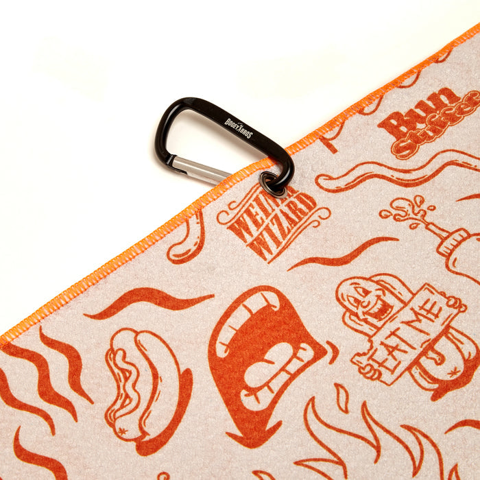 The Glizzy Gobbler - Towel is a beige microfiber towel with orange cartoon hot dogs, open mouths, and a figure holding an EAT ME sign. It features bold 2-sided print, orange stitching, a corner carabiner, and Weird Wizard Run Club branding.