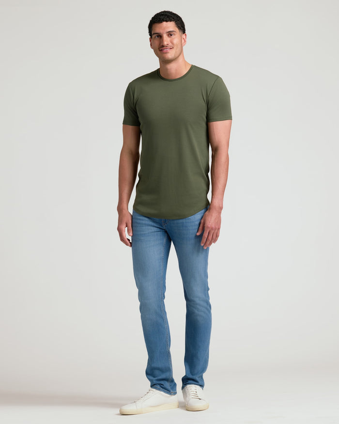 Man wearing an olive green Tall Pima Cotton Blend Curved Hem Crew 4-Pack t-shirt paired with light blue jeans and white sneakers, standing against a plain background.