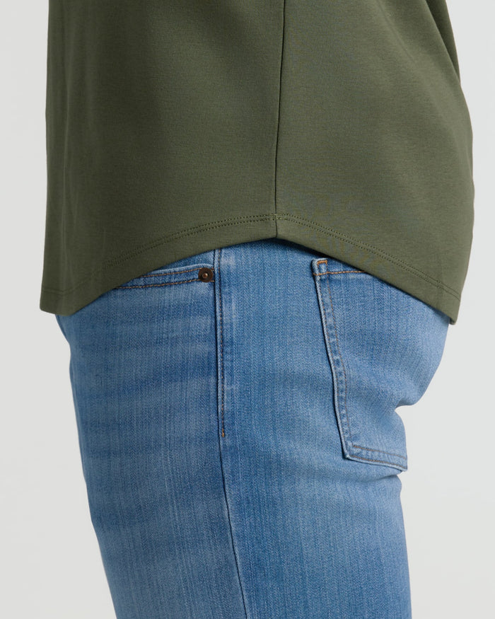 Side view of a person wearing light blue jeans and an olive green Grove Tall Pima Cotton Blend Curved Hem Crew, highlighting the curved hem detail against a plain background.