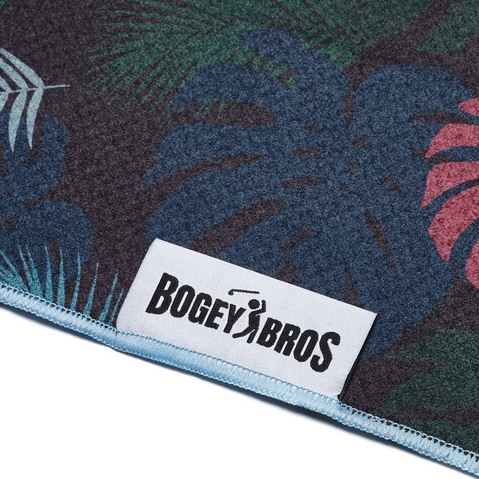 Close-up of the Haolie Nights - Towel, a dark microfiber towel with blue, green, and red tropical leaf patterns. Features light blue stitching, a corner clip and eyelet, and a white "Bogey Bros" label with a golfer silhouette.