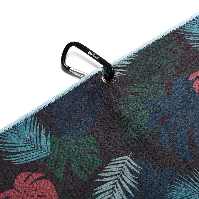 A close-up of the Haolie Nights - Towel shows its dark, tropical pattern with green, blue, and red leaves. A black carabiner clips to a metal eyelet at the light blue-edged corner. Enjoy vibrant 2-sided print design.