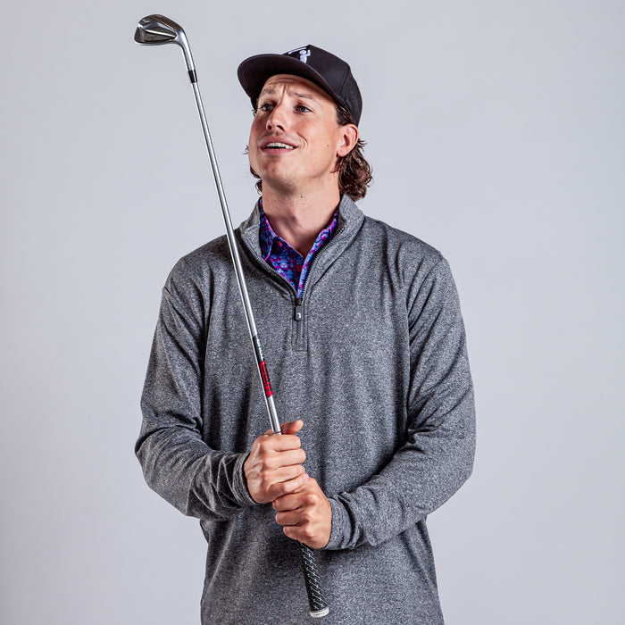 A man in a dark baseball cap and Heather Gray - Q-Zip holds a golf club upright, smirking as he looks up. Against a light gray background, his colorful patterned shirt peeks out beneath the athletic fit quarter-zip sweater.