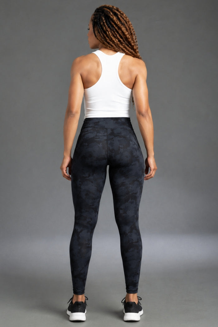 Rear view of a woman wearing high-waisted dark navy and black camouflage leggings with a white tank top, standing against a gray studio background. || Black Camo
