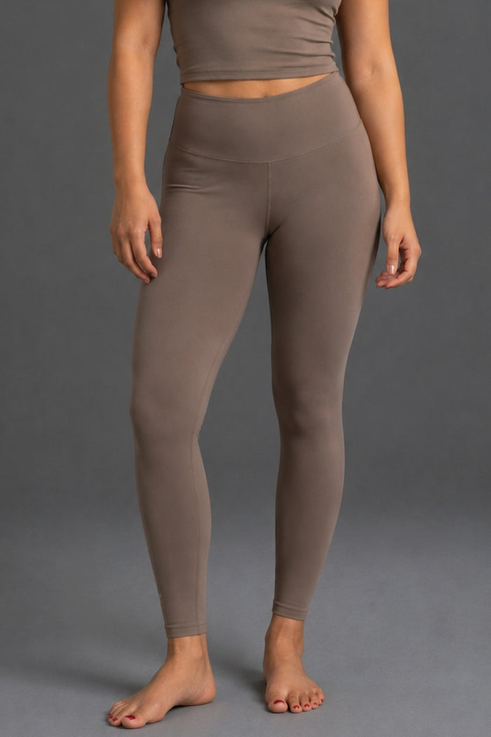 A model wearing high-waisted, ankle-length leggings in a solid taupe-brown color, shown from the waist down against a neutral gray studio background. || Mocha