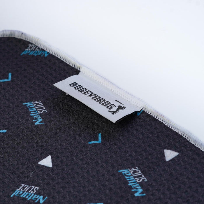 Close-up of the Natural Slice - Towel, a black microfiber towel with textured surface, blue and white text and triangle patterns. It features a white fabric tag with the black BOGEYBRO$ logo on the edge and 2-sided print.