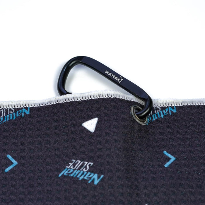 A black carabiner with BOESHIELD in white is attached to the Natural Slice - Towel, a dark microfiber towel with blue and white arrow patterns and "Natural Slice" printed on both sides for style and visibility.
