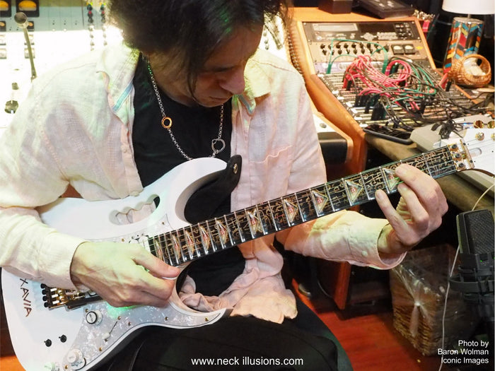 image of steve vai playing a white electric guitar in a recording studio.  there is a fret protector on the guitar. It is black with light brown symbols all over it. Every other fret has a brown, black and greenish triangle on it with a white steve vai logo in the center of it. The steve vai logo makes the word "vai" with an upside down triangle, a right side up one, and a line going across the triangles with a curl at the end next to the triangle that is upright.
