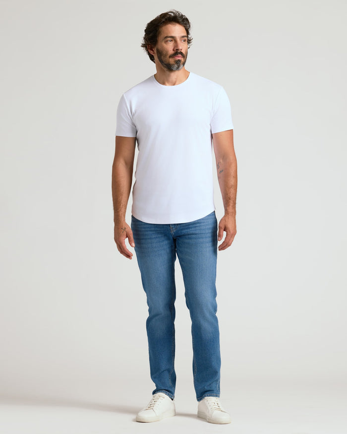 Male model wearing a white Pima Cotton Blend Curved Hem Crew 7-Pack t-shirt paired with blue jeans and white sneakers, standing against a plain background.