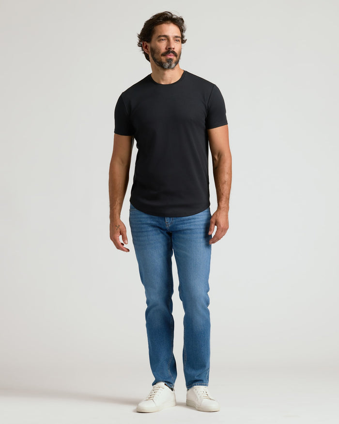 A man wearing a black Pima Cotton Blend Curved Hem Crew 4-Pack shirt paired with blue jeans and white sneakers, standing against a plain light background.