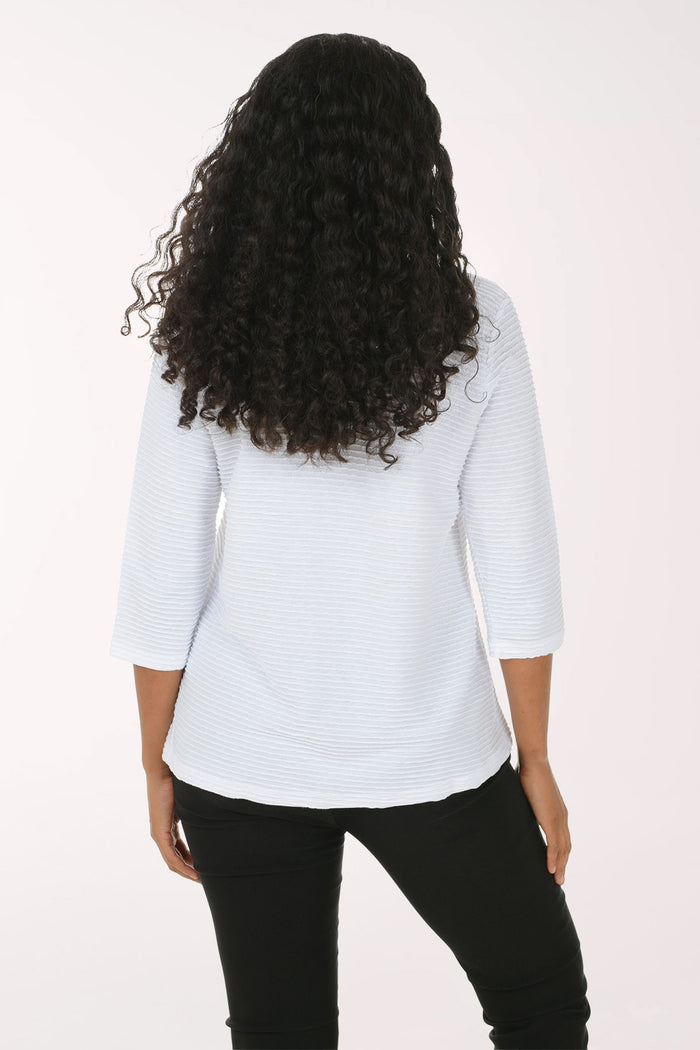 A woman with long curly black hair wearing a white textured three-quarter sleeve top and black pants, posing against a plain white studio background.