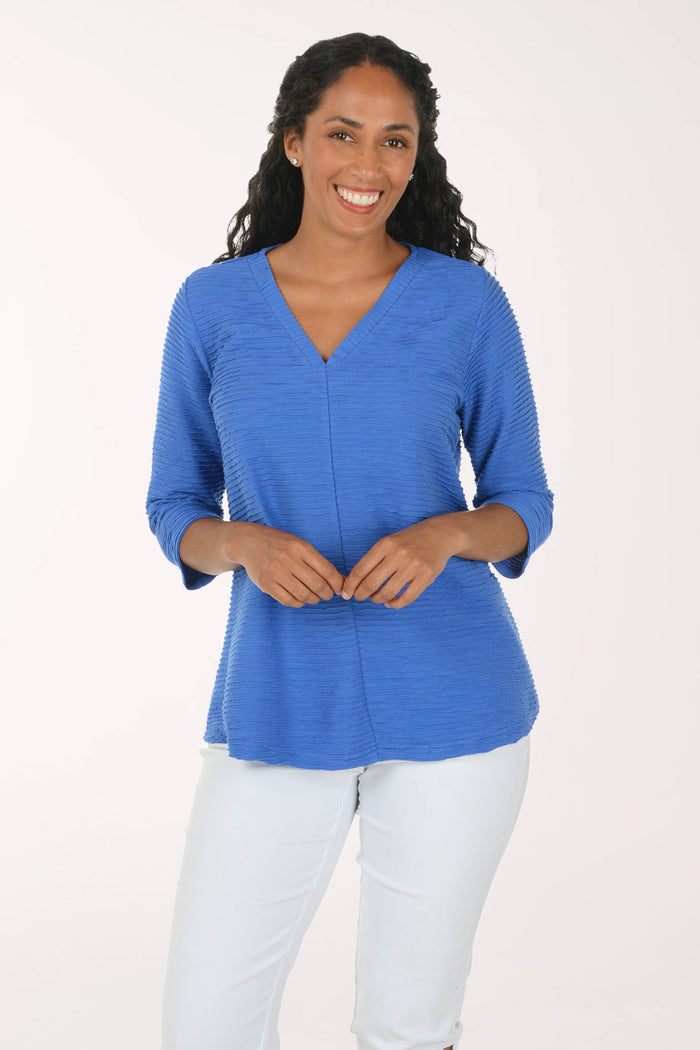 A woman modeling a blue ribbed knit top with three-quarter sleeves and a V-neckline, paired with white pants, against a plain white studio background.