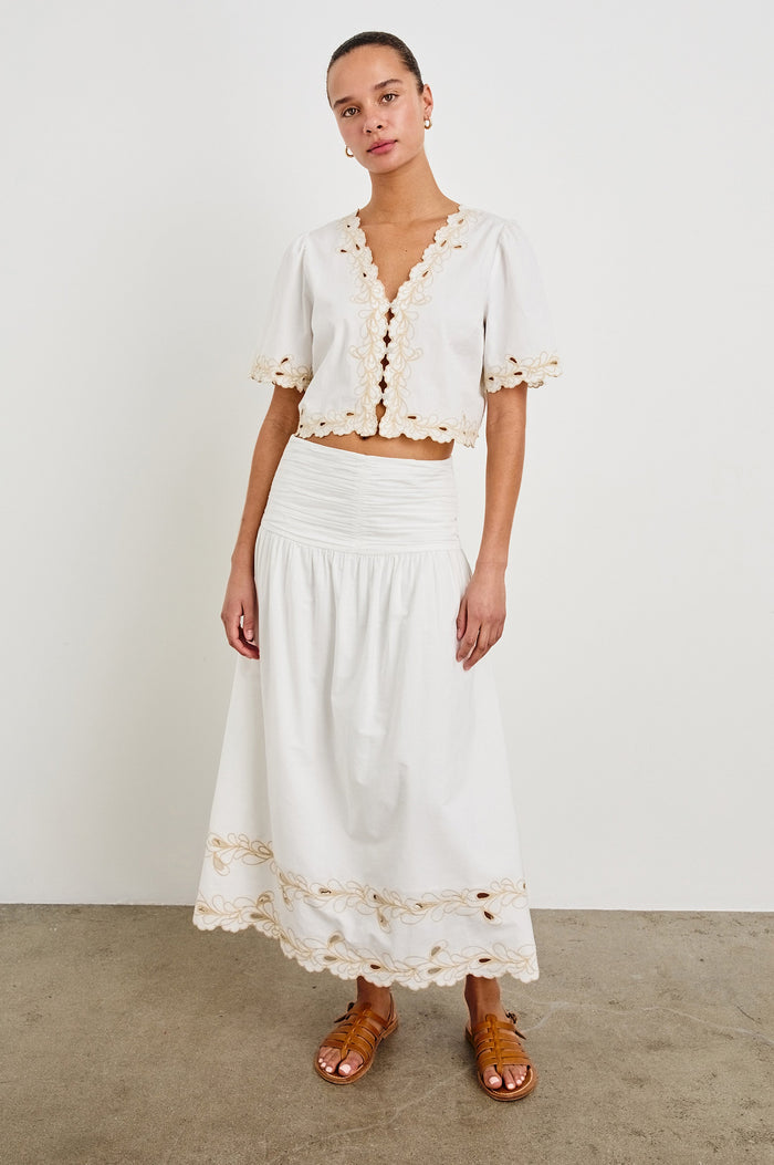 Front image of model wearing a white, cotton poplin midi skirt with a lightly gathered waist that cinches and drops at the hips. Details include beige embroidered eyelets at the hem. Worn with brown sandals and matching cropped top.
