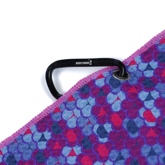 A close-up of the Whale Tail - Towel, a 2-sided microfiber towel with blue, purple, and pink scale patterns. A black BOGEY BROS carabiner is attached to the edge via clip and eyelet, with purple stitching along the border.