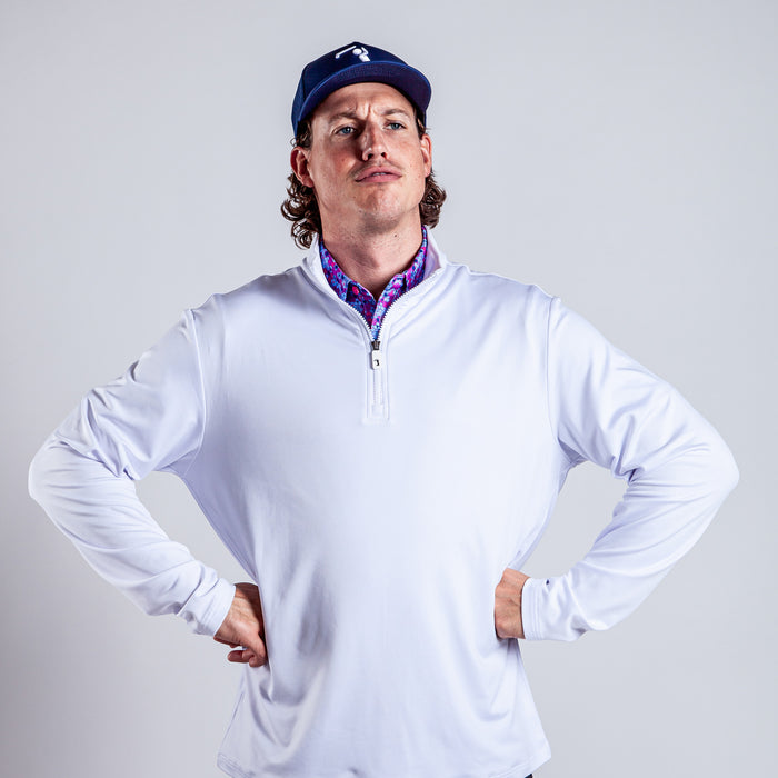 A man with curly hair and a serious, confident expression stands against a light gray background, wearing the White - Q-Zip athletic fit long-sleeve shirt over a purple patterned layer and a blue baseball cap.
