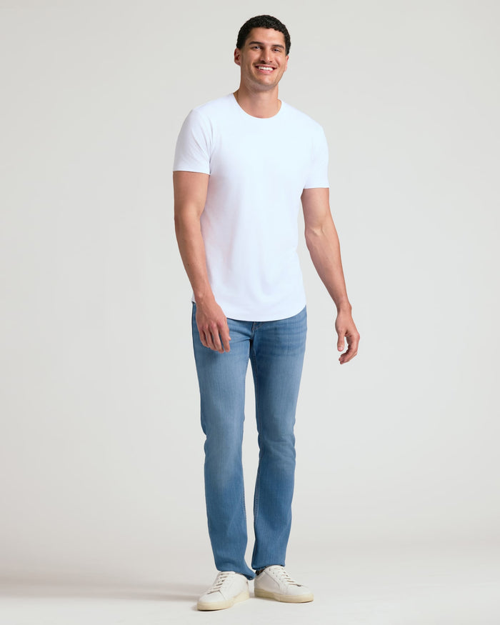 A smiling man wearing a white Tall Pima Cotton Blend Curved Hem Crew 4-Pack T-shirt paired with light blue jeans and white sneakers, standing against a plain background.